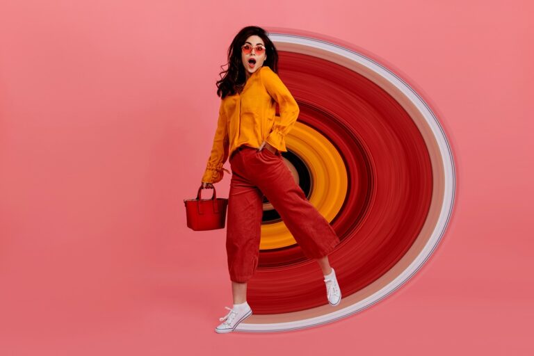 Perky girl in stylish glasses stares into camera in amazement, walking on pink background. Brunette in culottes and orange blouse posing with red handbag